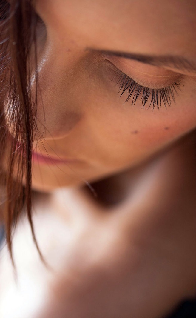 La imagen muestra un primer plano artístico de una mujer con los ojos cerrados y una expresión serena. Su piel tiene una textura natural y luminosa, con pequeños lunares visibles. La iluminación resalta los contornos de su rostro, dando una sensación de calidez e intimidad. Un mechón de cabello cae suavemente sobre su cara, aportando un toque orgánico y desenfadado a la composición. La imagen transmite una sensación de calma y bienestar, ideal para representar el autocuidado y la belleza natural.