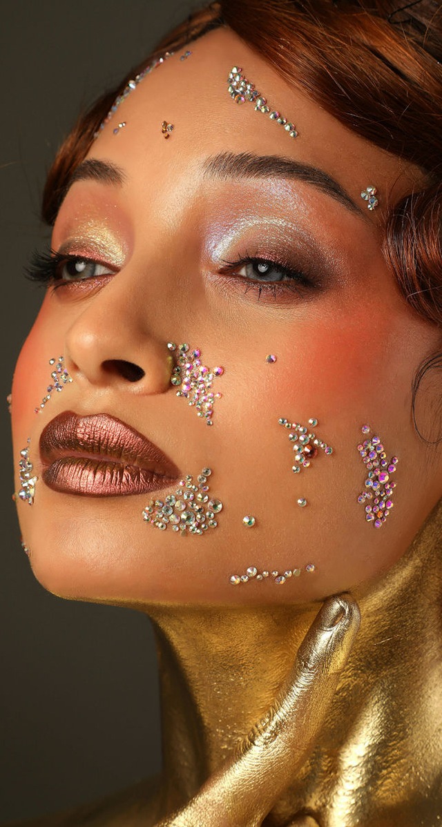 Esta imagen presenta un retrato de gran impacto visual y sofisticación. La modelo luce un maquillaje artístico con sombras en tonos metálicos y labios en un deslumbrante tono bronce, que aportan un aire futurista y lujoso. Su piel está adornada con cristales brillantes estratégicamente dispuestos sobre su rostro, creando un efecto de luz y dimensión.  Su cuello y mano están completamente cubiertos en un baño dorado, reforzando la estética de opulencia y poder. Su mirada intensa y expresión serena transmiten una mezcla de confianza y misticismo. El peinado con ondas y mechones estructurados aporta un toque vintage con un aire moderno.  Los tonos dorados y metálicos contrastan elegantemente con la iluminación suave y el fondo neutro, resaltando la riqueza de los detalles. Esta imagen encarna la fusión entre el arte y la belleza, evocando sofisticación, glamour y vanguardia.