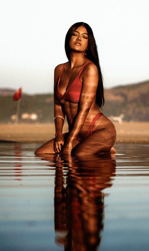 Esta imagen transmite sensualidad, fuerza y conexión con la naturaleza. Una mujer con un bikini rojo intenso posa de rodillas en aguas poco profundas, reflejando su silueta en la superficie del agua con una composición visualmente impactante.  Su piel, bañada por la luz cálida del atardecer, brilla con un tono dorado que resalta su figura atlética y esculpida. Su expresión serena y segura, con los ojos entrecerrados, evoca una sensación de empoderamiento y plenitud.  El fondo difuminado con montañas y una bandera roja aporta profundidad y un ambiente de aventura y libertad. La simetría entre la modelo y su reflejo en el agua añade un toque artístico a la imagen, reforzando la armonía y el equilibrio de la composición.  Esta fotografía encapsula la esencia de la belleza natural, el poder femenino y la conexión con el entorno en un instante de calma y confianza absoluta.