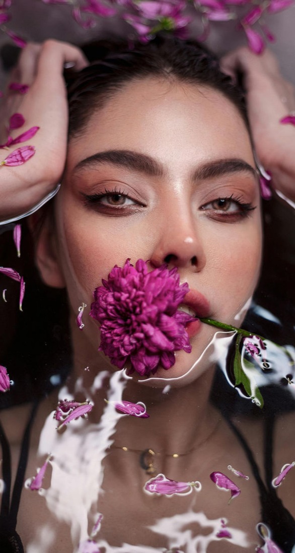 Esta imagen es una obra de arte visual que combina la belleza, la feminidad y la naturaleza en una composición etérea y sofisticada. Una mujer flota en el agua con una flor fucsia entre sus labios, rodeada de pétalos dispersos que realzan la armonía del entorno.  Su mirada intensa y enigmática, enmarcada por cejas perfectamente definidas y un maquillaje en tonos cálidos, transmite serenidad y magnetismo. La piel húmeda y luminosa refleja la suavidad del agua, resaltando su textura impecable y la frescura del momento.  La combinación del reflejo del agua y los pétalos flotantes crea una atmósfera casi onírica, evocando sensaciones de relajación y conexión con la naturaleza. Los tonos rosas y morados aportan un aire romántico y poético, mientras que el contraste con su piel y la transparencia del agua añade profundidad y misterio.  Esta imagen encapsula la fusión perfecta entre el arte, la belleza y el bienestar, transmitiendo una sensación de calma, lujo y sofisticación.