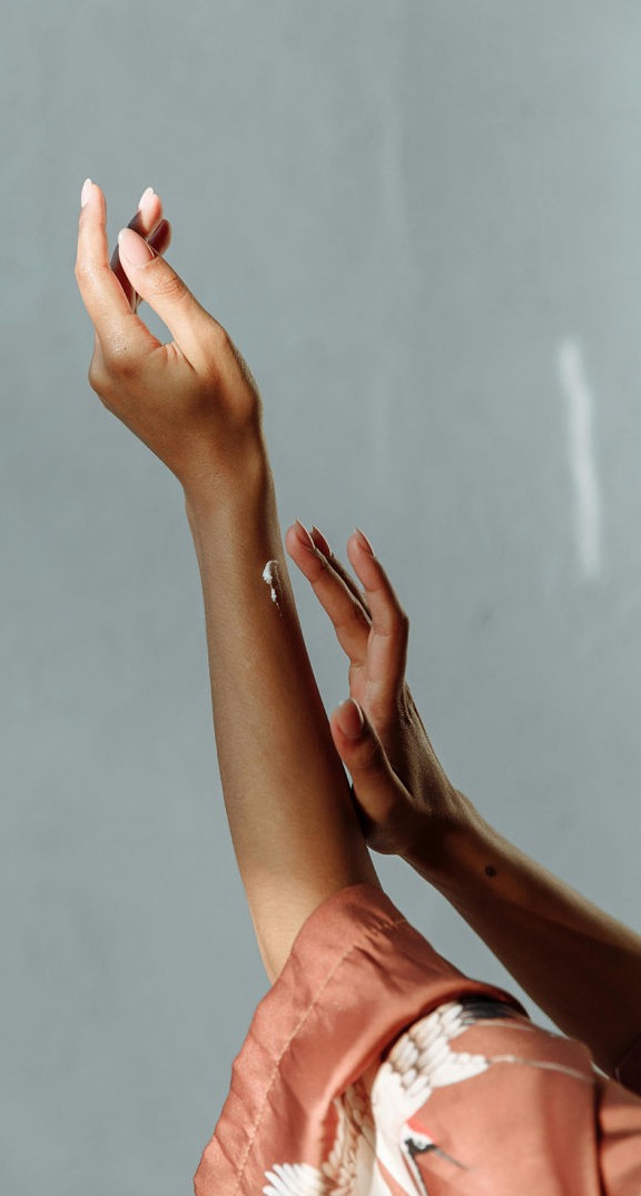 Esta imagen captura un momento de autocuidado y bienestar. Se observan unas manos femeninas aplicando una pequeña cantidad de crema en la piel del antebrazo, en un gesto de hidratación y mimo personal. La luz natural resalta la suavidad y el brillo de la piel, mientras que el fondo neutro aporta elegancia y simplicidad a la escena.  El kimono en tonos tierra con detalles de grullas añade un toque sofisticado y relajante, evocando una atmósfera de calma y ritual de belleza. La postura de las manos, con las uñas cuidadas y el movimiento delicado, enfatiza la sensación de conexión con uno mismo y la importancia del cuidado de la piel en la rutina diaria.