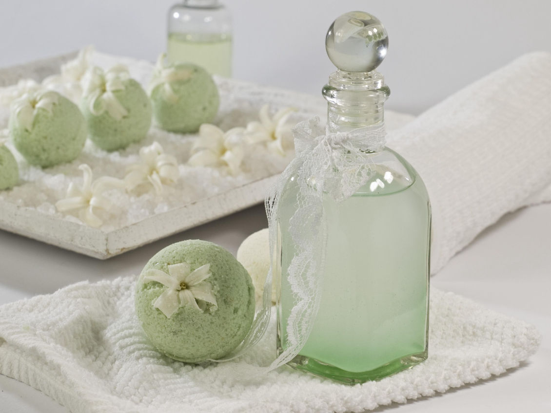 Esta imagen transmite una sensación de lujo y bienestar, mostrando productos de baño y cuidado personal en tonos verdes suaves y blancos. Las bombas de baño decoradas con lazos y el frasco de vidrio con encaje refuerzan una estética elegante y natural, ideal para representar productos de aromaterapia, spa o cosmética ecológica.