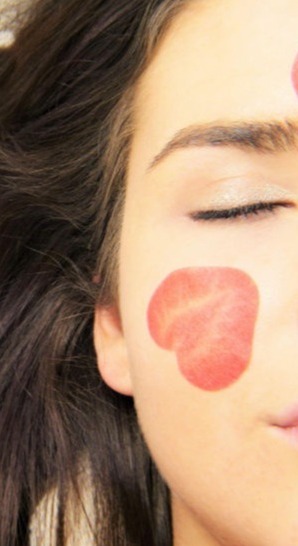 La imagen muestra un primer plano de una mujer con la piel luminosa y relajada. En su mejilla hay un pétalo de rosa con forma de corazón, lo que sugiere una rutina de cuidado de la piel con ingredientes naturales o un tratamiento rejuvenecedor. Su ojo está cerrado, lo que transmite una sensación de calma y bienestar. El fondo desenfocado y la iluminación cálida refuerzan la idea de un ambiente spa o de relajación.