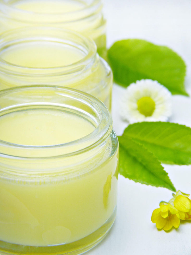 Esta imagen representa cremas naturales, posiblemente elaboradas con ingredientes botánicos. Los tarros de vidrio con una textura cremosa de tono amarillo suave, acompañados de hojas verdes y flores blancas y amarillas, refuerzan la idea de cosmética ecológica y orgánica. Es ideal para transmitir un mensaje de productos naturales y sostenibles en la industria del cuidado de la piel.