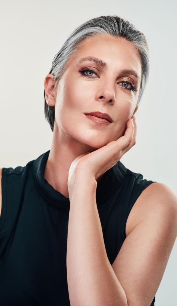 Esta imagen presenta un retrato impactante y evocador de una mujer con una mirada penetrante e intensa. La luz dorada del atardecer baña su rostro, creando un juego de sombras y reflejos que resaltan la profundidad de sus ojos azules y la sensualidad de sus labios.  Su cabello rubio cae estratégicamente sobre parte de su rostro, aportando un aire de misterio y sofisticación. La iluminación dramática y la textura suave de su piel crean un contraste cautivador, mientras que su postura y expresión sugieren confianza y una elegancia innata.  El tono cálido de la imagen, sumado a su atuendo negro, refuerza una estética moderna y cinematográfica. Este retrato captura la belleza en su faceta más artística y enigmática, evocando sensaciones de poder, magnetismo y autenticidad.